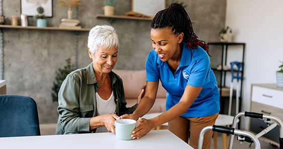 A Visiting Angels caregiver happily offers a hot cup of coffee to a senior woman with a walker, seated at her kitchen table.