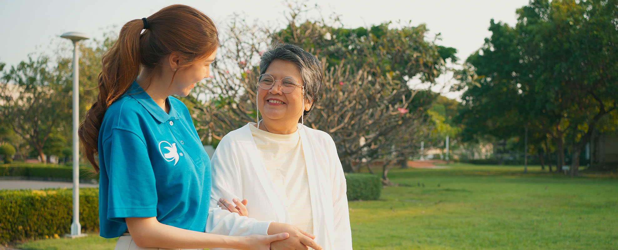 A female senior home care provider from Visiting Angels holds the arm of a smiling elderly woman as they take a walk in the park.