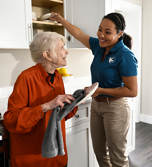 A female Visiting Angels caregiver helps an elderly woman clean and put away the dishes.